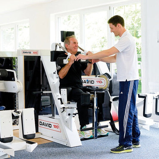 Medizinische Trainingstherapie: Trainer unterstützt Senior bei Übung am DAVID Gerät während MTT-Training im ZAR Heilbronn.